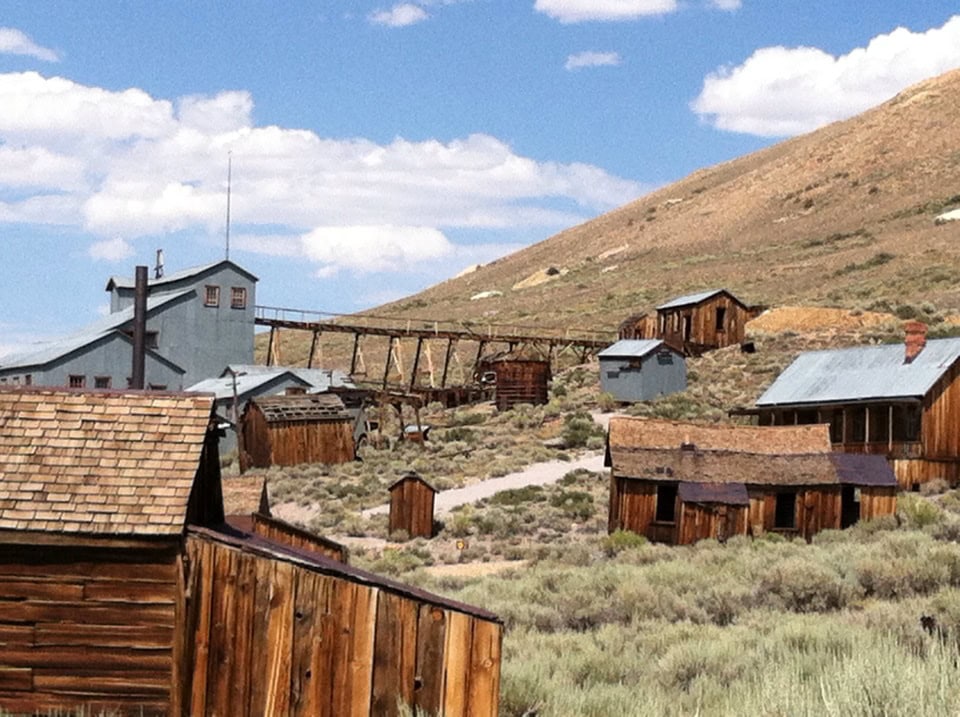 Rustic frontier ghost town with old wooden buildings and hills under blue sky, evoking a vintage western farm or mining town setting.