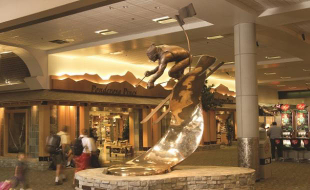 Bronze sculpture of a boy surfing in a shopping mall atrium with storefronts and shoppers in the background.