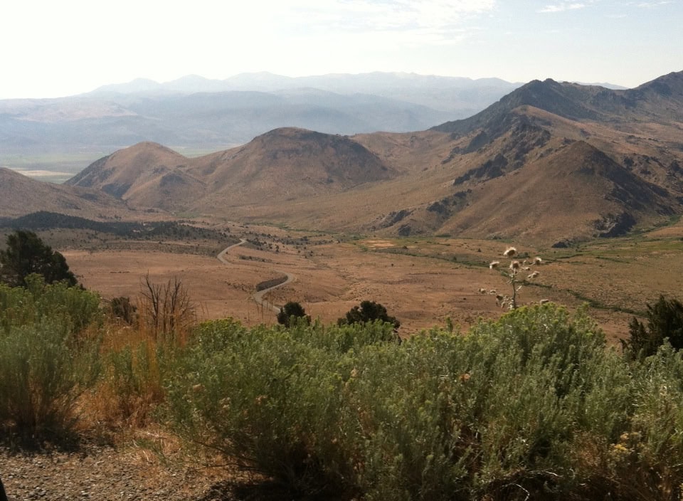 Vast mountain landscape with rolling hills, dry plains, sparse vegetation, and a winding road under a partly cloudy sky.