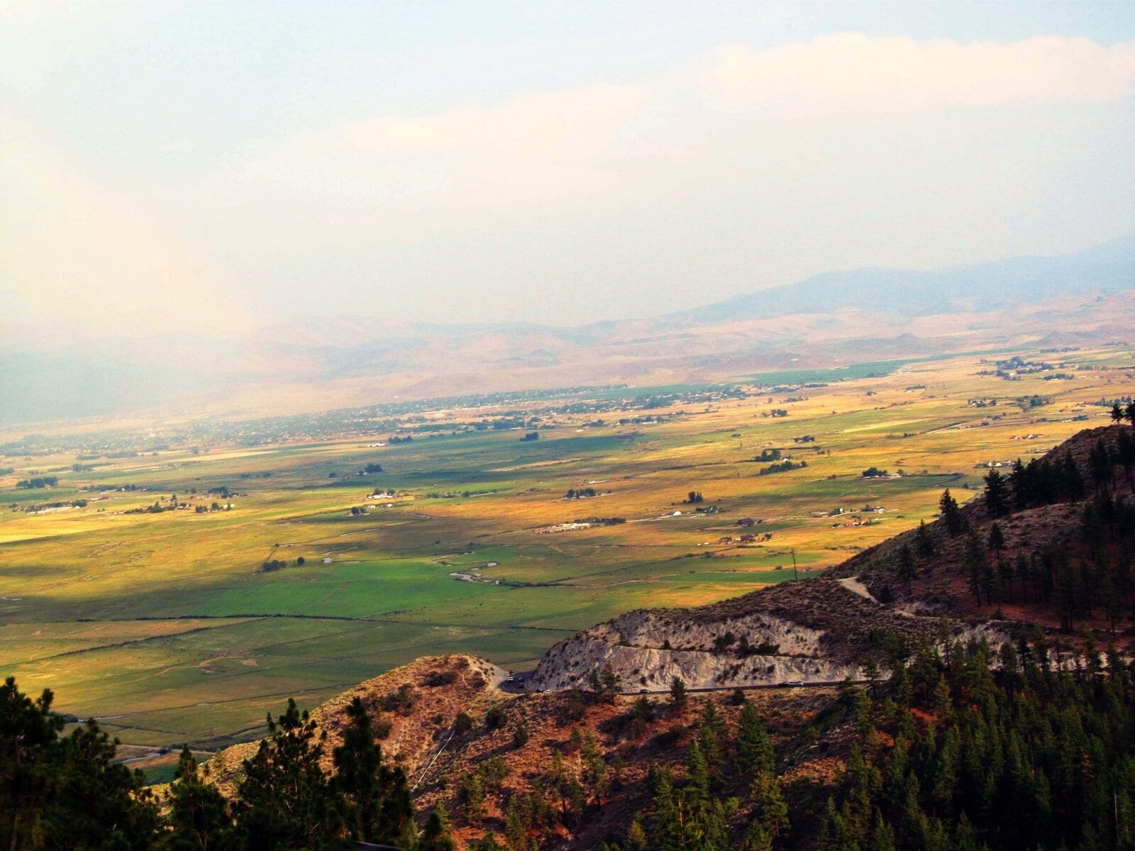 Vast agricultural valley seen from a mountain, with patches of green fields, scattered farms, rolling hills, and distant mountains under a partly cloudy sky.