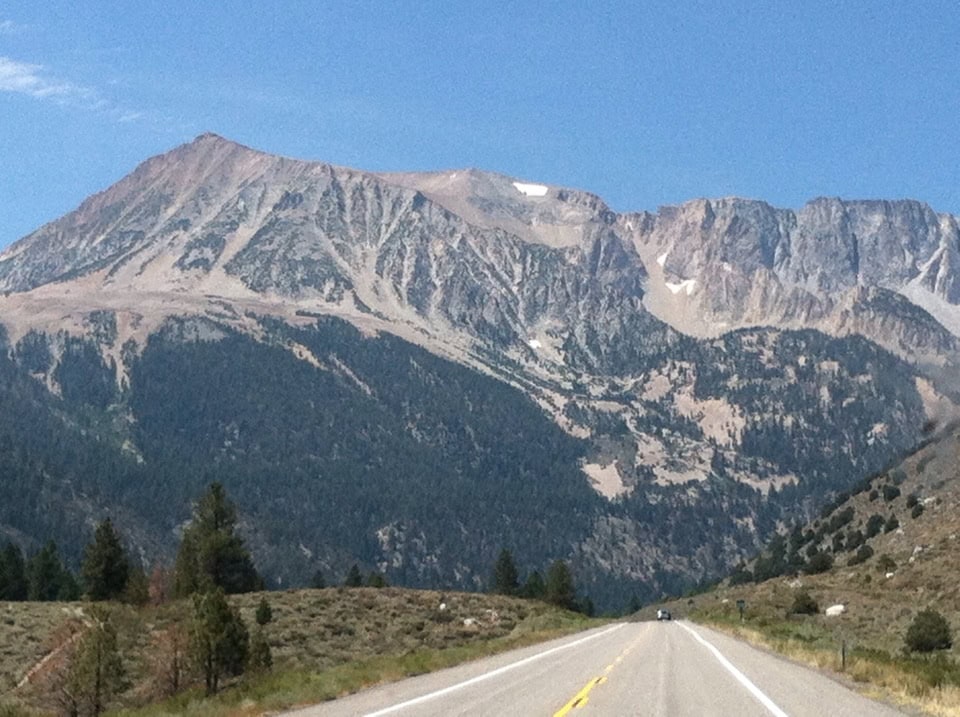 Vast mountain landscape with a scenic open road in the foreground, lush greenery, and rugged peaks under a clear blue sky, perfect for travel and adventure photography.