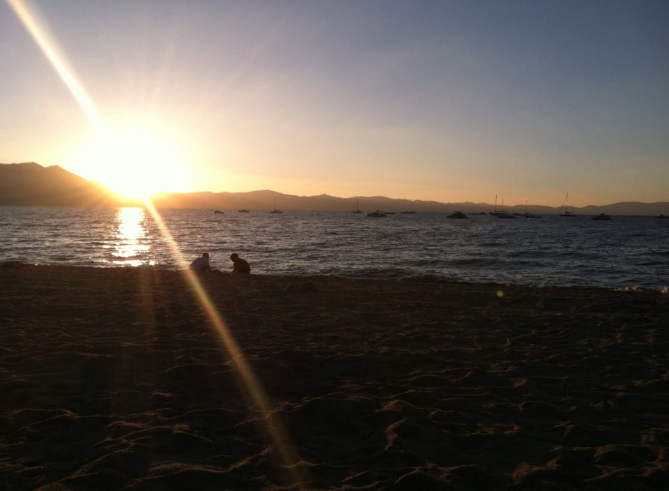 Sunset over calm ocean with silhouettes of two people on sandy beach and sailboats in distance, creating a peaceful, romantic seaside scene.