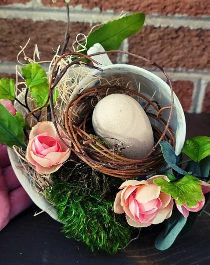 Egg nestled in a bird's nest surrounded by pink flowers, green foliage, and natural textures, creating a charming and rustic spring or Easter-themed decorative arrangement emphasizing nature and renewal.