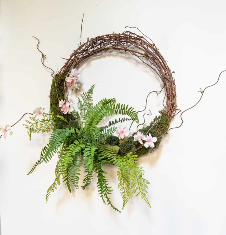 Vine and fern floral wreath with soft pink flowers, moss, and twigs on a white background, perfect for spring or rustic decor.