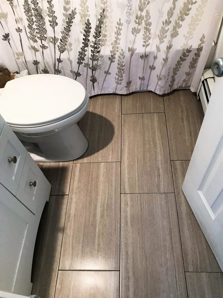Lavender and beige bathroom tiles with shower curtain featuring floral pattern, white toilet, and small white vanity. Perfect for cozy, rustic bathroom decor.