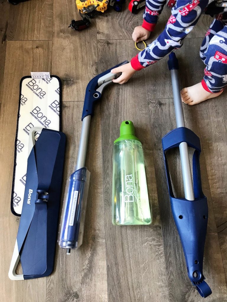 Child sweeping wooden floor with cleaning tools and water bottle.