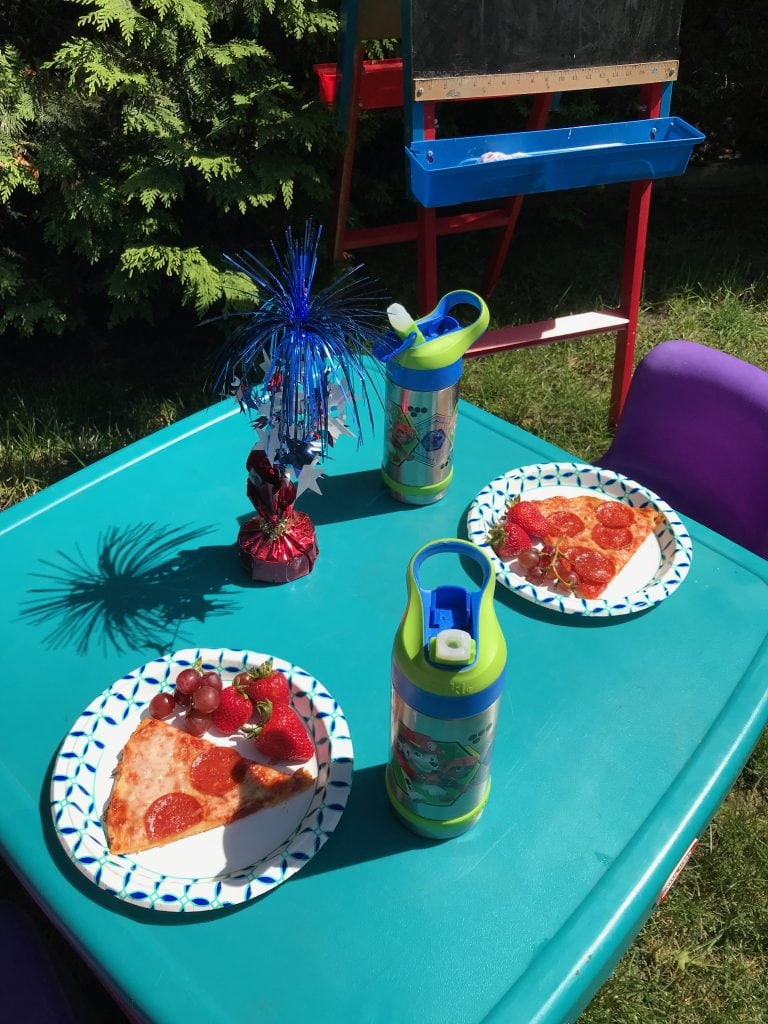 Colorful outdoor kids' birthday party setup with pizza, strawberries, grapes, and themed water bottles on a bright turquoise table.