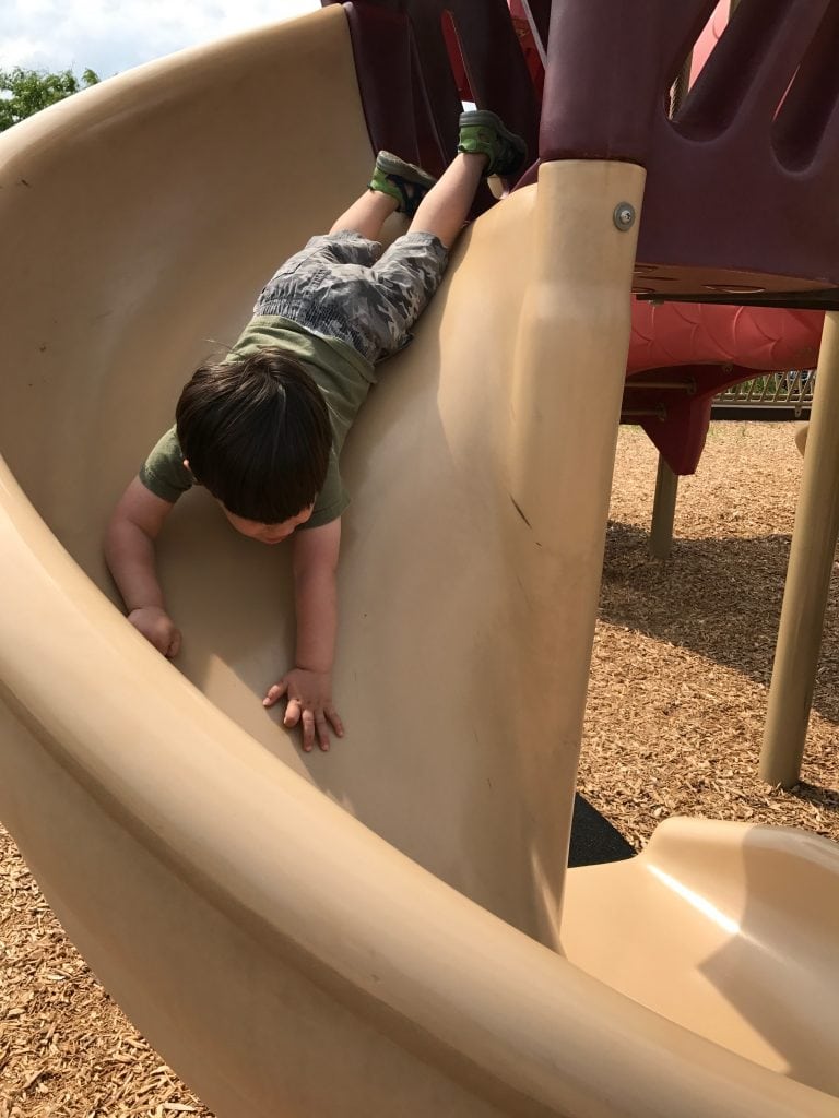 Child playing on a playground slide, wearing camouflage shorts and a green t-shirt, enjoying outdoor activities and playground fun.