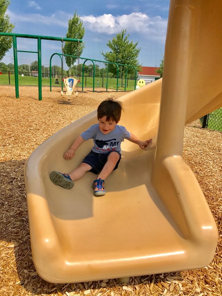 Bright young boy playing on beige playground slide at park, kids slide and swings, outdoor playground equipment, family fun, childhood joy, sunny day, green trees, blue sky.