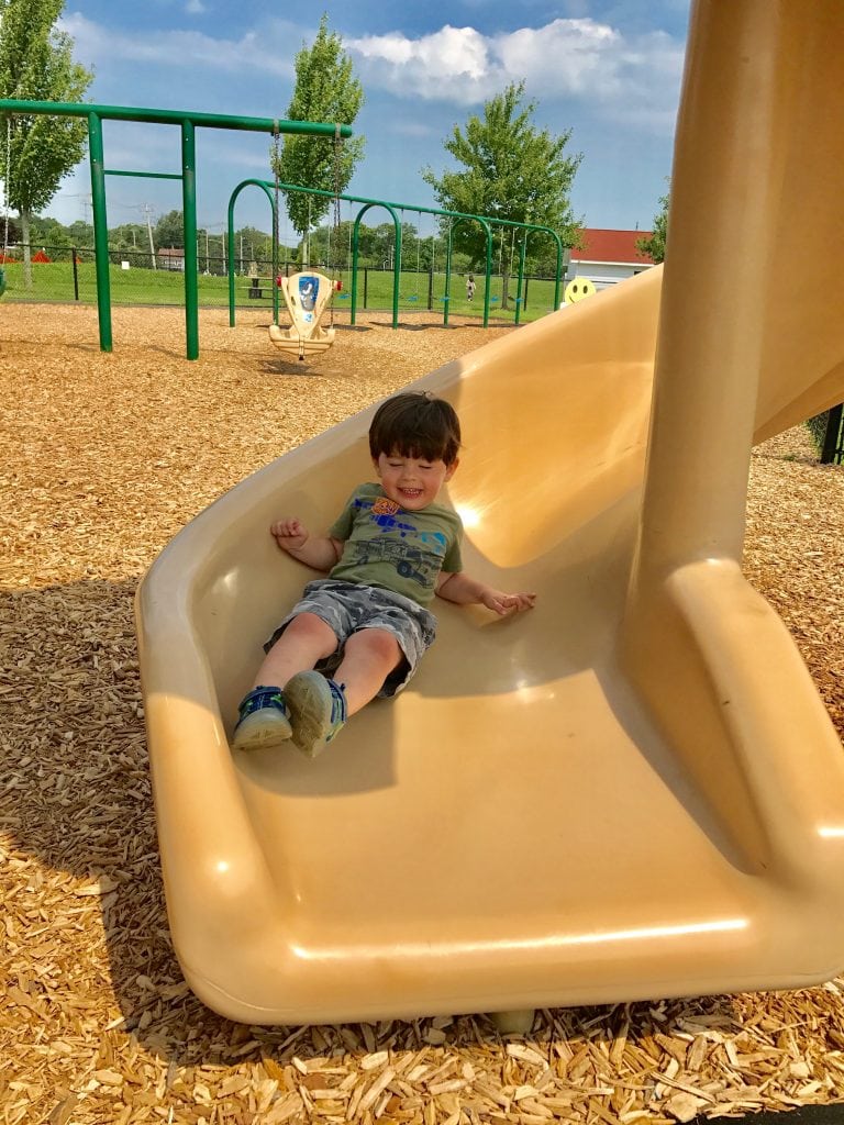 Happy young boy sliding down a beige playground slide at a park with wood chip ground cover and green trees in the background.