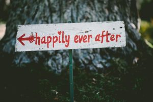 Whimsical outdoor wooden sign with red hand-painted lettering that says "happily ever after" and an arrow, set against a natural background for romantic photography and wedding decor.