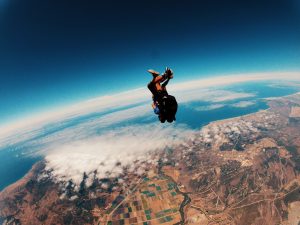 Skydive over landscape with patchwork farmland and ocean view, adventurous skydiver free-falling in high-altitude jump, thrilling experience, extreme sports, aerial photography, breathtaking scenery.