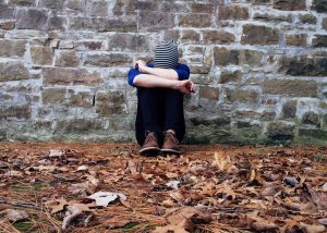 Sad child hugging knees against stone wall, autumn leaves on ground.