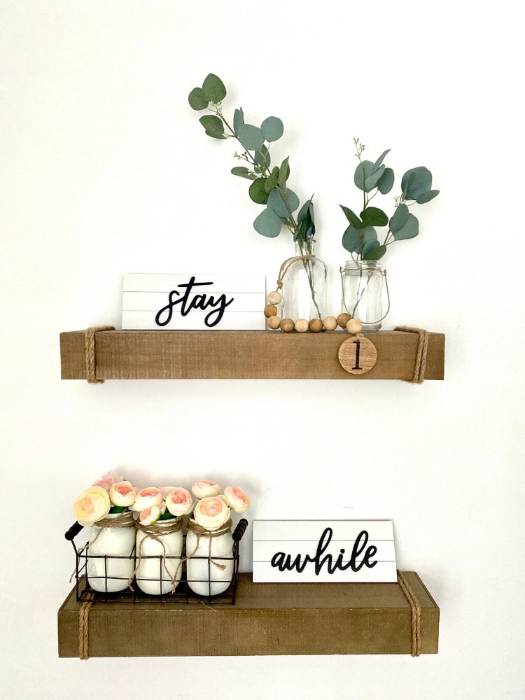 Eucalyptus branches in glass jars with "stay" and "awhile" signs on rustic wooden shelves, modern minimalist home decor for a cozy living space.