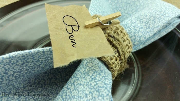 Delicate blue fabric napkin wrapped with twine and a “Bev” hand-written note clip on a glass jar table setting, perfect for personalized dining or special occasions.