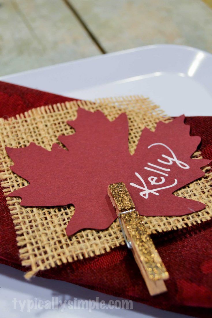 Rust-colored maple leaf with "Kelly" written in white script, attached to gold glitter clothespin, on a textured burlap and red fabric background, perfect for fall or Thanksgiving decor.