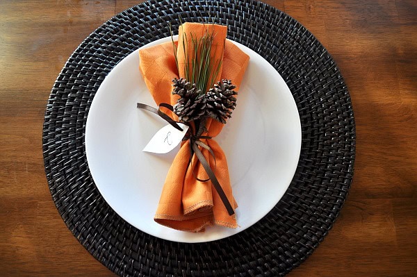 Elegant table setting with a black woven charger, white plate, orange napkin, pinecones, and greenery for holiday or festive dining experience.