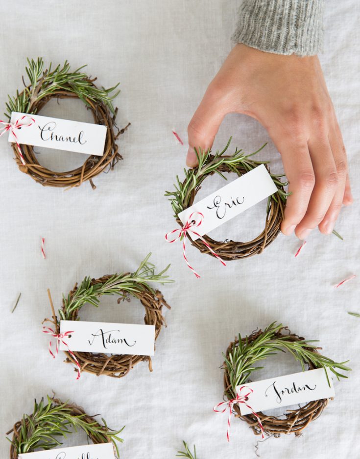Hand arranging small DIY Christmas wreaths with sprigs of fresh rosemary, personalized with name tags, for holiday home decor and festive gift ideas.