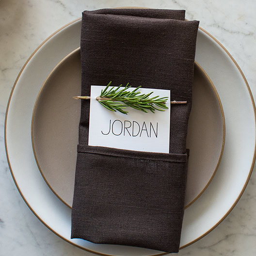 Elegant place setting with dark napkin, personalized name card, and fresh rosemary on a stylish white and beige plate, ideal for special occasions or wedding tablescapes.