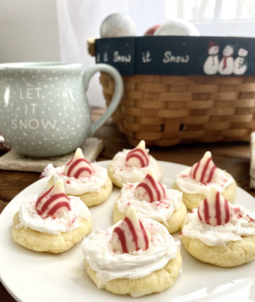 Hershey's Candy Cane Hug North Pole Cake Mix Christmas cookie on plate, cookie with white frosting, coconut, candy cane bits, plated with Christmas coffee mug and basket