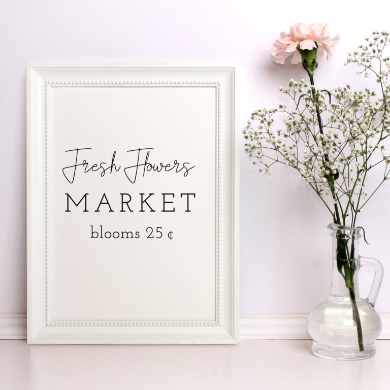 Delicate pink carnations and white baby's breath flowers arranged in a glass vase next to a white framed chalkboard sign with floral market announcement.