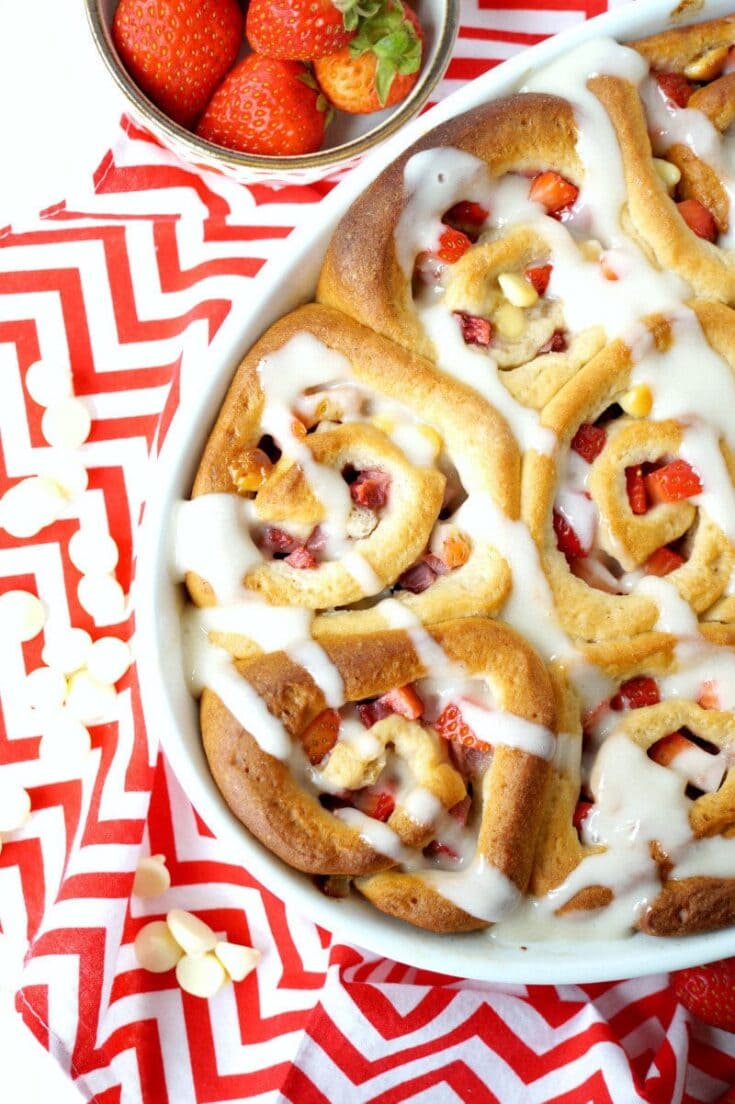 Fresh strawberry cinnamon rolls drizzled with icing, served with a side of ripe strawberries on a red and white patterned cloth.