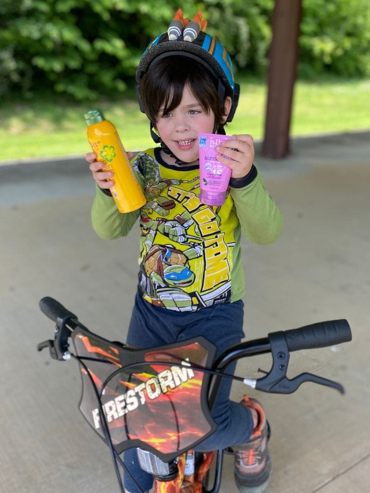 Brightly dressed young boy with a superhero-themed shirt and bike helmet holding sunscreen and lotion, outdoors on a sunny day, emphasizing family summer skincare and active kids.