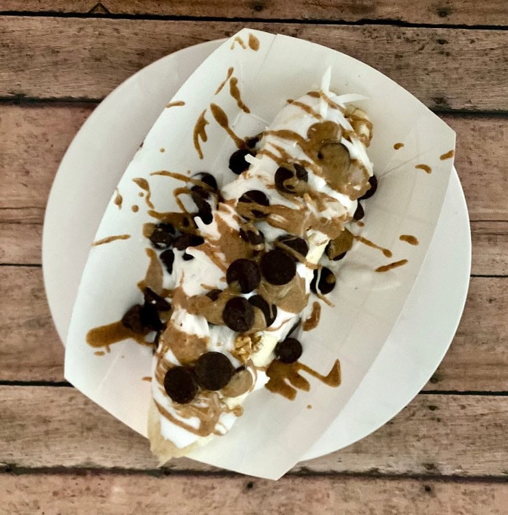 Sweet banana split with chocolate chips, whipped cream, caramel drizzle, and chocolate sauce on a white plate, served on a rustic wooden table, perfect for dessert lovers.