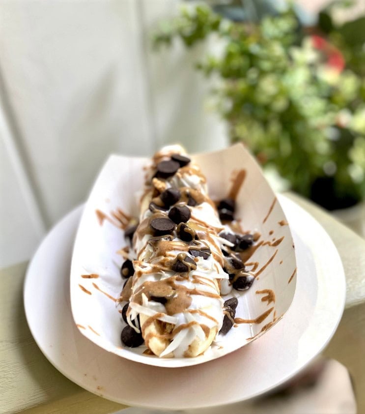 Banana split with chocolate chips, whipped cream, caramel sauce, and shredded coconut on a white paper liner, showcasing delicious toppings and fresh ingredients at A Hundred Affections.