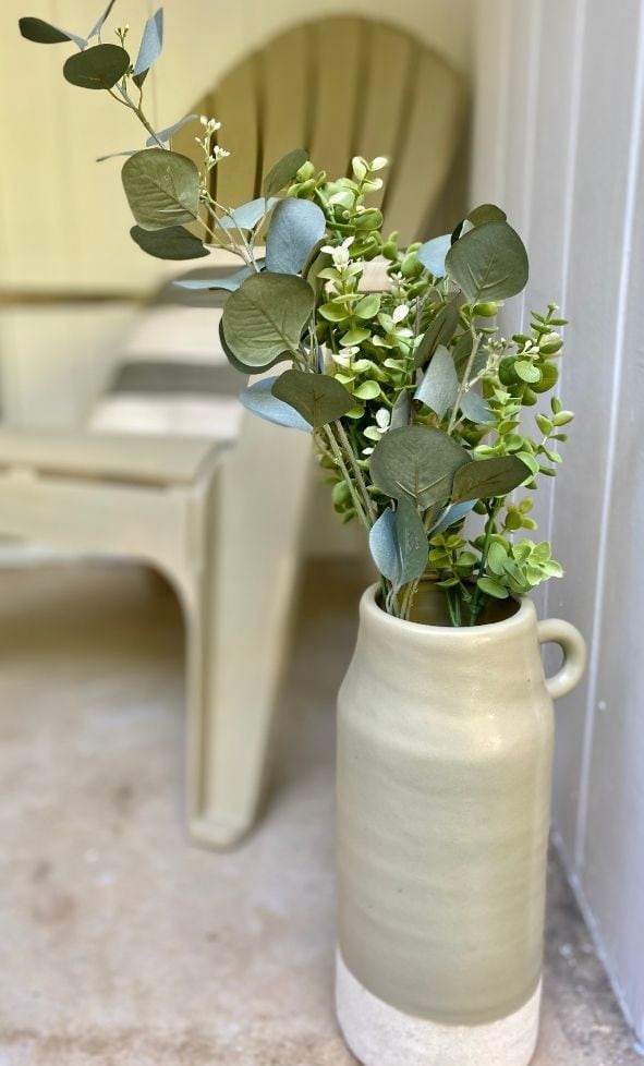 Lush green eucalyptus and succulents arrangement in a tall cream ceramic vase on a rustic floor with a cozy seating area in the background.