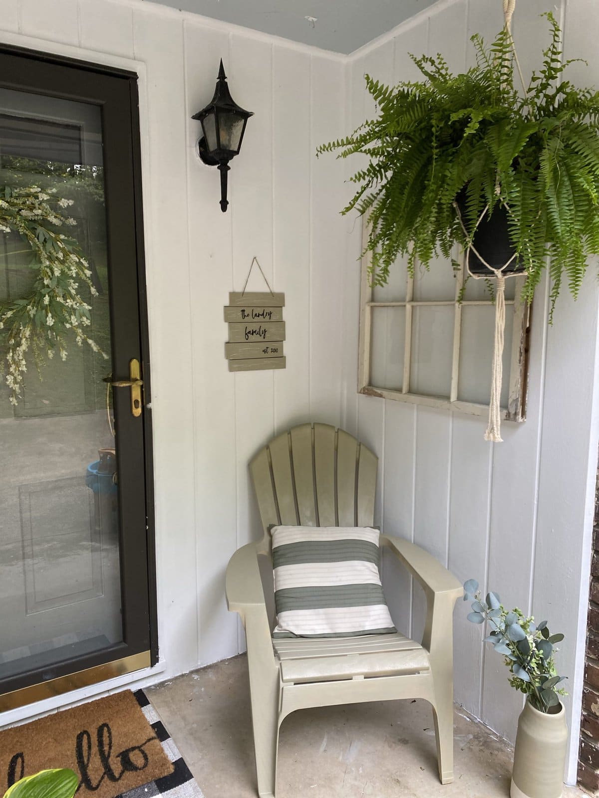 Cozy porch corner with white wooden chair, striped pillow, hanging fern plant, decorative wall window, and welcoming "hello" doormat, perfect for outdoor relaxation and charming home decor.