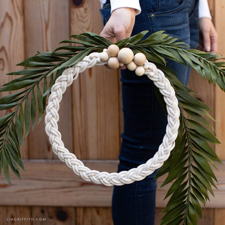 Handcrafted floral wreath with white braided rope, wooden beads, and lush green foliage, ideal for home decor or special occasions.