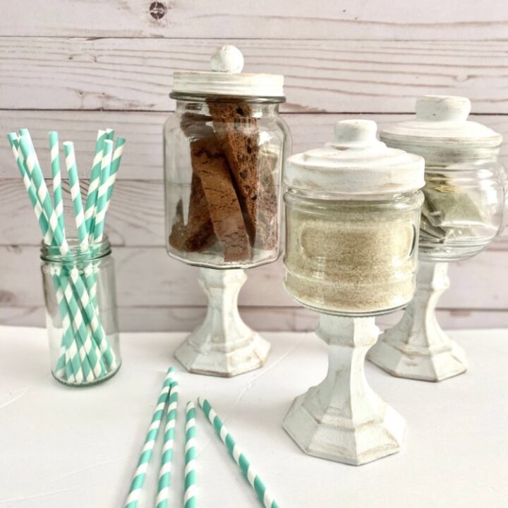 Deluxe vintage glass jars with white wooden bases holding assorted baked goods and sugar, turquoise striped paper straws, and a rustic white background perfect for a charming dessert display.