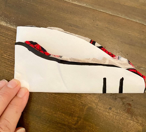 A handmade fabric shoe craft with red and black plaid details, partially cut out from paper or fabric, on a wooden surface for DIY sewing or craft project.