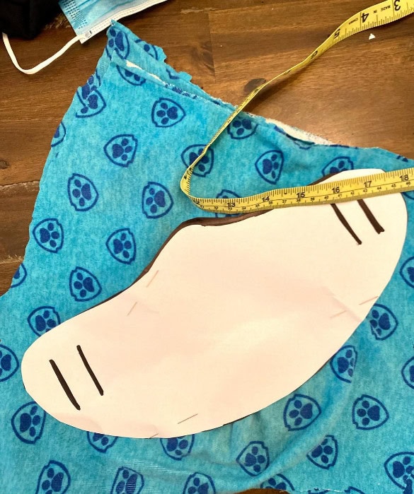 Soft blue fabric with paw print pattern, cut-out mask template, measuring tape, and sewing supplies for custom pet-themed mask creation.