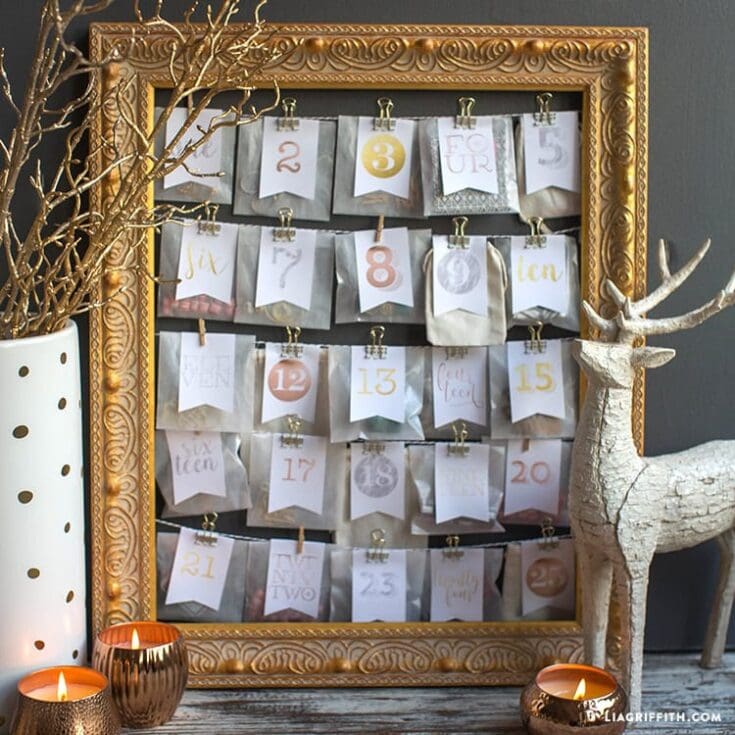 Advent calendar display with gold frame, featuring numbered paper bags clipped to strings, surrounded by seasonal decor like candles, a white reindeer figure, and a white vase with dried branches, perfect for holiday home decor.