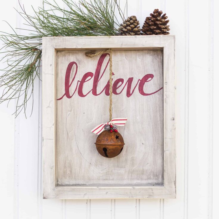 Festive holiday decoration featuring a rustic wooden frame with the word 'believe' in red, a jingle bell hanging with a ribbon, pinecones, and pine branches for Christmas cheer.