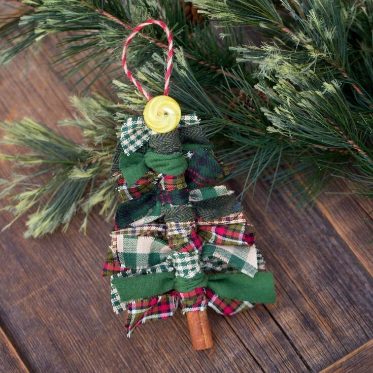 Handmade fabric bow Christmas tree ornament with plaid patterns, green ribbons, a yellow button, and a red-and-white twine loop, resting on pine branches for holiday decor.