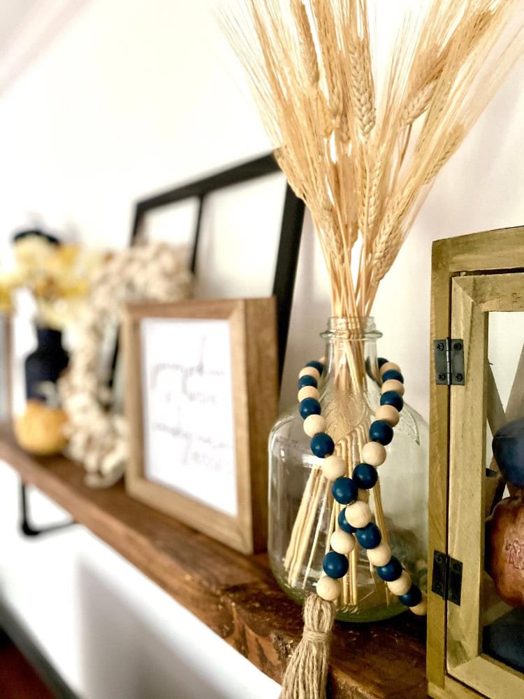 Wheat bouquet with beaded tassel and decorative accents on a wooden shelf in home decor setting.