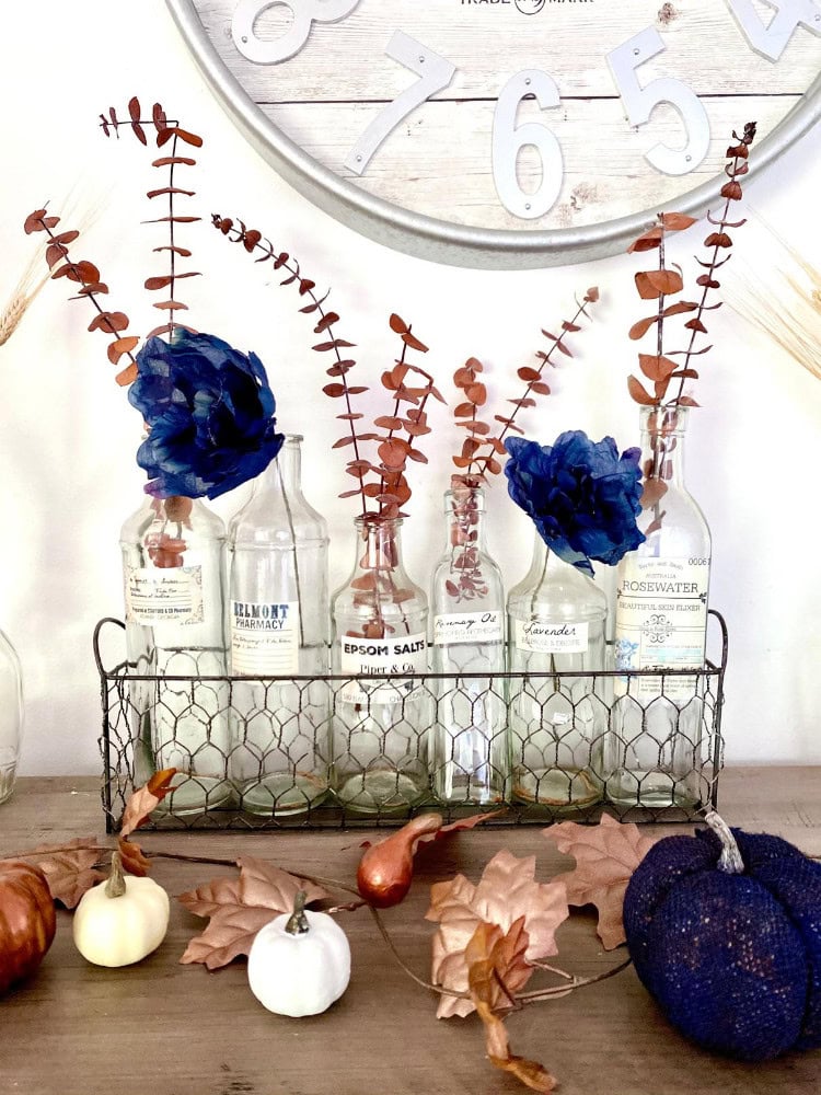 Eucalyptus and blue hydrangea flowers in glass bottles, rustic decor with pumpkins, autumn leaves, and a wall clock in a farmhouse style for cozy fall decoration.