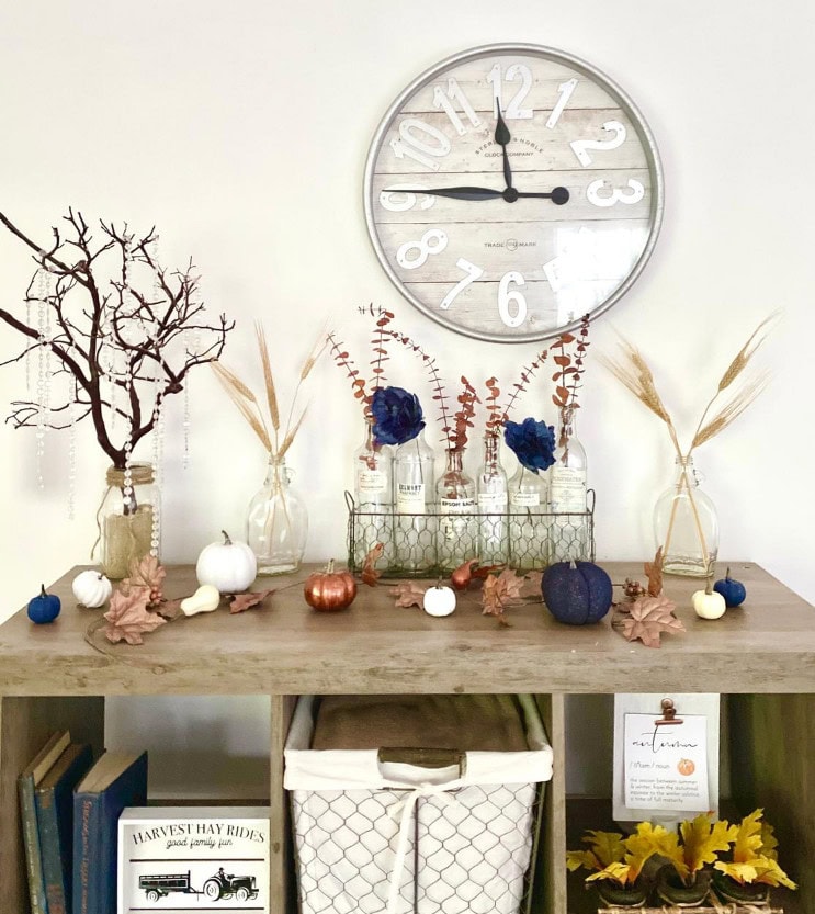 - Elegant fall home decor featuring white pumpkins, dried leaves, and glass vases with dried flowers on a wooden table against a white wall with a modern wall clock.