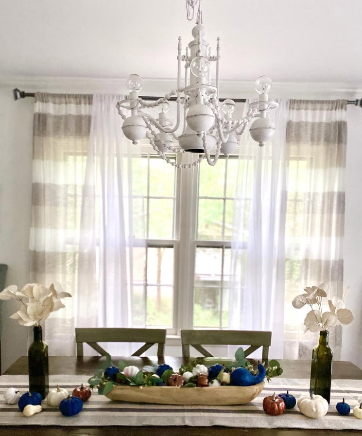 Pumpkin fall centerpiece with white and blue pumpkins on wooden dining table decorated with flowers and eucalyptus, white chandelier and sheer curtains behind, cozy autumn home decor.