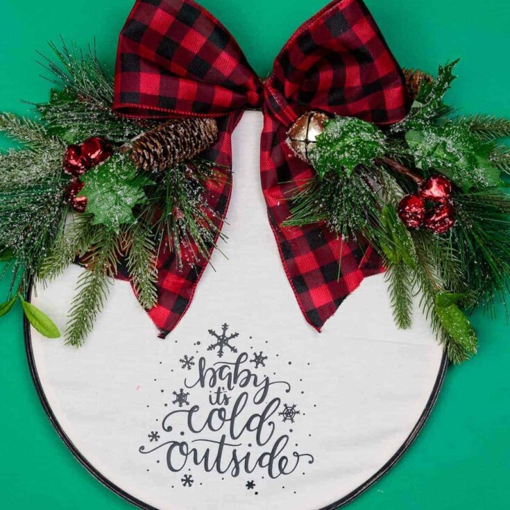 Red and black buffalo plaid bow with holiday greenery, pinecones, holly, and red berries on a white circular holiday sign that says "happy it's cold outside" for Christmas decor.
