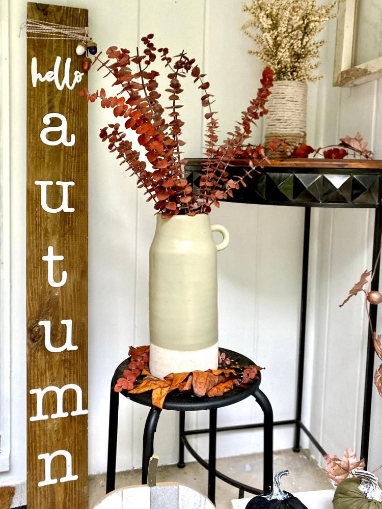 Hello autumn with a rustic wooden "hello autumn" sign, dried reddish-brown eucalyptus in a tall beige ceramic vase on a black metal stool, surrounded by fall leaves, cozy decor, and seasonal accents for autumn decor, curated by A Hundred Affections.