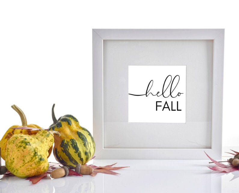 Colorful decorative gourds with fall leaves and a framed "hello fall" sign on a white background, perfect for autumn home decor or seasonal greeting ideas.