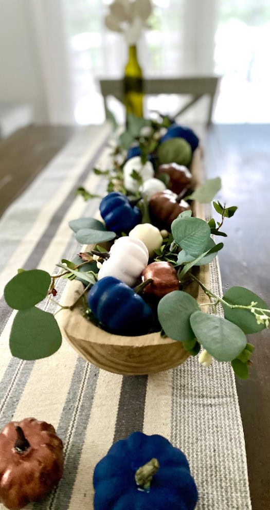 Colorful painted pumpkins and eucalyptus greenery arranged in a rustic wooden tray for fall decor or centerpiece. Ideal for autumn tablescapes, harvest celebrations, and seasonal decorating.