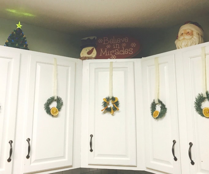 Christmas wreaths hanging on white kitchen cabinets with holiday decor and a "Believe in Miracles" sign in the background.