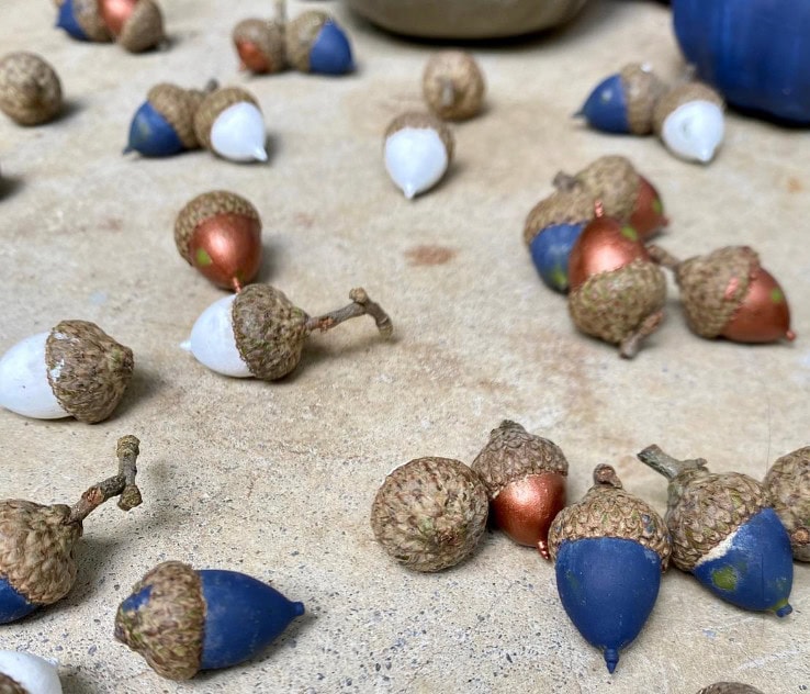 Colorful acorns with painted caps scattered on a sandy surface for fall or craft decor.