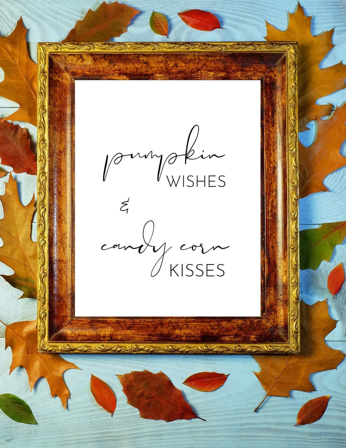 Cozy autumn scene with a framed quote reading "Pumpkin wishes & candy corn kisses," surrounded by colorful fall leaves on a light blue wooden surface, perfect for seasonal holiday decor.