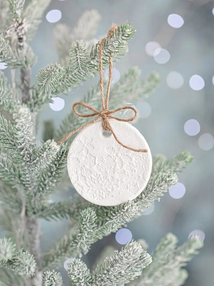 Elegant white Christmas ornament with embossed snowflake design, hanging on a snow-dusted pine tree branch, creating a festive holiday atmosphere.